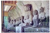 Color photograph of a row of ca. ten seated stone statues in front of a rock. Three-quarter view. One of the statues is about twice as large as the others.