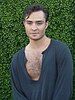 A Caucasian man dressed in a blue low cut shirt standing in front of a green hedge