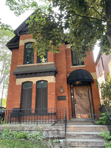 A two-storey brown brick building in the Summertime with a tree covering part of it.