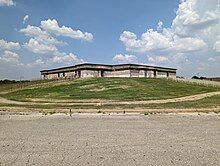 A two story tan stucco building. The first floor is covered by an earthen berm. The stucco is being repaired.