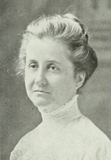 A middle-aged white woman, her hair dressed in a bouffant updo, wearing a high-collared white lacy blouse or dress