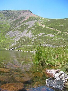 A clear lake in the foreground with a steep mountain ridge beyond