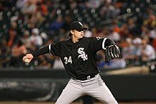 A young man in a black baseball jersey and cap with white trim and white baseball pants throws a baseball with his left hand.