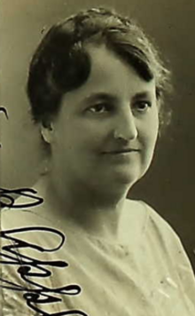 A middle -aged white woman, dark hair braided across the crown, wearing a simple light-colored top; part of her signature is visible