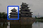 A 3-storied castle tower with black wooden walls located on a platform of unhewn stones above a water filled moat. The tower is connected to a lower structure on one sides.