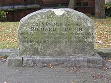 A stone gravestone with a curved top, with several lines of inscription. Trees, grass, and a wall are visible in the distance.