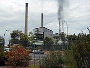 Four chimneys, two emitting strong black smoke, from industrial building with Western Power logo above text "Wester Power Muja Power Station", seen through wire fence across parking aisle.