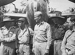 Five men in light-coloured uniforms standing in front of a military aircraft