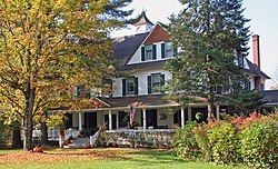 A large house with peaked roofs behind two trees. There is an American flag flying from the front porch and pumpkins around the walk.