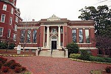 A one-story brick building with grey concrete stairs in the center leading to a door with a column on either side of it. There are three long windows on each side of the building.