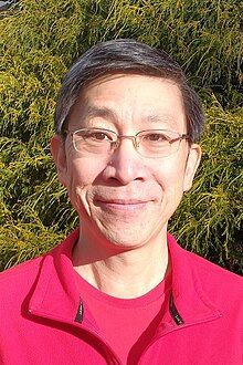 Headshot of an Asian-American man with short salt and pepper hair and glasses, wearing a read shirt and jacket, standing in front of a chartreuse evergreen bush