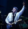 A man wearing a black tie and white dress shirt with an electric guitar hanging from a strap around his neck. His left arm is raised, with his index finger pointed upwards. In the background is a drum set.