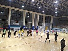 This image depicts two teams playing futsal on an indoor wooden court.