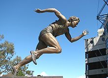 bronze statue of a woman moving from a crouched running position into an all out spring. Statute us on a black pedestal. It is located outside against a clear blue sky.