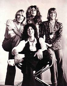 A posed black-and-white shot of four men. Three are standing behind a sitting man. The first standing man, at left, has long fair to blond hair. His right leg is raised and he clutches his arms, which are resting on his knee. The second standing man is looking to his right, he has long, wavy hair and wears a floral shirt. The third standing man has fair hair, shorter than the previous. He wears a floral jacket, a tie without a shirt, and rests his hands on the back of the chair in front of him. The sitting man has dark, curling hair, which is just over shoulder length. His left leg is resting on his right knee and he grabs it with both hands. He wears a white shirt, a dark vest and dark pants. The chair appears to be a deck chair with metal legs and frame. The background is grey.