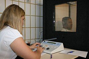 a female medical professional is seated in front of a special sound-proof booth with a glass window, controlling diagnostic test equipment. Inside the booth a middle aged man can be seen wearing headphones and is looking straight ahead of himself, not at the tester, and appears to be concentrating on hearing something