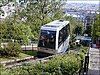 A photograph looking down the line. In the foreground is a cabin with its distinctive half-glass construction. The lower station is in the middle ground and a panorama of Paris fills the background.