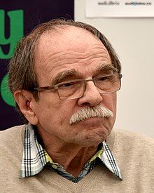 close-up of Jaroslav Uhlíř wearing a checkered shirt under a tan sweater, looking left of camera with pursed lips