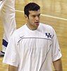 A man in his twenties with dark hair wearing a white warm-up jacket with the letters UK in blue on the left shoulder and a blue Nike "swoosh" on the right shoulder