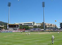 A ground with a bowler running in to bowl, with stands and floodlights in the background