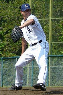 A man in a white baseball uniform, blue baseball cap, and black baseball glove pitching a ball right handed.