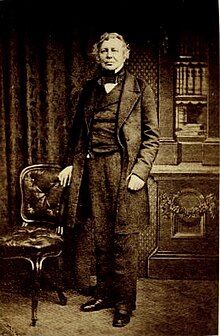 Standing studio portrait of Effingham Wilson, formally dressed in cravat, waistcoat and long jacket, between a chair and a bookcase