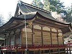 Three-quarter view of a proportionally tall wooden building with a veranda with red hand rail. The corner of the eaves appear cut off.