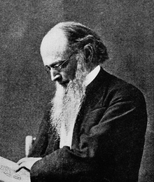 Head and shoulders photograph of a man in an overcoat. He has a full beard with sideburns, has a shaved upper lip and is balding. He wears a serious expression and is looking down at a paper with an insignia drawn on it.