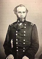 Sepia toned photo shows a man with a salt-and-pepper beard. He wears a dark military uniform with two rows of buttons.