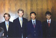 Four men in black suits with bluish-white dress shirts and brightly coloured ties standing in front of a wall composed of wooden panels.