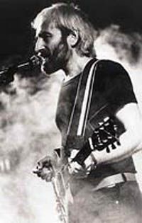 black-and-white image of Dežo Ursiny playing guitar and singing into a microphone onstage, surrounded by smoke