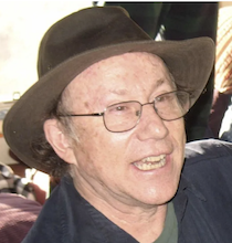 small headshot of Holladay a white man in glasses wearing a brown hat and a blue shirt