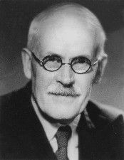 Black and white portrait photograph of Sir Harold Jeffreys looking into the camera. He is wearing a shirt, tie and jacket. He has a moustache and is wearing spectacles.