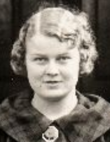 A young white woman with curly blonde hair, wearing a plaid dress with a pin at the throat