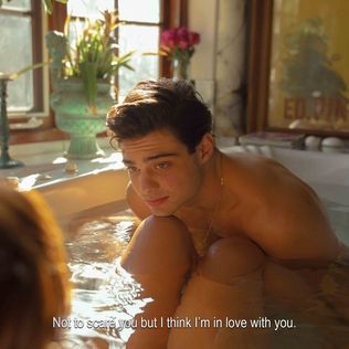 A photograph of a man with short, dark hair sitting upright in a bathtub full of water, leaning forward against the knees of a woman who is also in the tub. The woman's back is to the camera and only her legs and the back of her head are in frame. The caption on the photograph reads "Not to scare you but I think I'm in love with you."