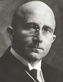 Bald, elderly man with gold-rimmed spectacles, wing collar, and serious expression