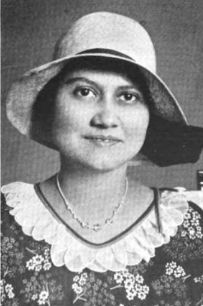 A smiling woman with olive skin and dark hair, wearing a hat with a wide brim, and a floral print dress or blouse with a ruffled white collar.