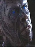 A closeup of a man's head; his face is covered in tattoos, and his eyes are glowing blue.