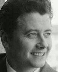 Publicity shot of smiling, clean-shaven man in suit and tie, with swept-back, bouffant hairstyle.