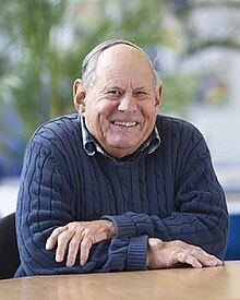 A white man in old age, seated at a conference table, smiling broadly at the camera. He wears a thick, navy-blue woollen jumper.