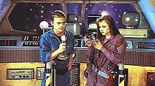 Two teenagers, one boy and one girl, on the bridge of an untidy space station