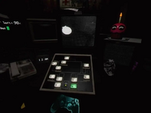 A screenshot of an office desk. On top of the desk is a computer monitor controlled by various buttons, which projects a black-and-white camera feed. To the left of the monitor is a landline phone and to the right is a restaurant prop that looks like a cupcake with eyes