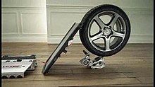 a flat board strikes the tread of a car tyre at a 45-degree angle. The tyre is balanced on top of a smaller piece of machinery on a wooden floor.