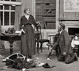 Standing woman in drawing room with bearded man kneeling amid debris