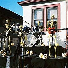 Four golden skeletons in a rock band are playing drums, guitars, and singing in a backyard