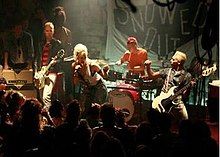 A band consisting of three men and a woman perform onstage in a crowded club in front of a large black sign with "Snowed Out" written in white. The woman stands in the center and leans forward while singing into a microphone. Two guitarists flank her, and the drummer plays behind her.