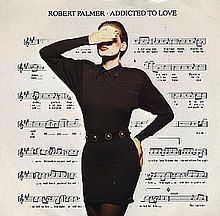 A model with a hand raised covering her eyes, her other hand is on her hip. She is wearing a black dress and dark tights. Her face is heavy with makeup, the white of her face contrasting dramatically with her red lips. The background is a filled by a sheet of music notation.