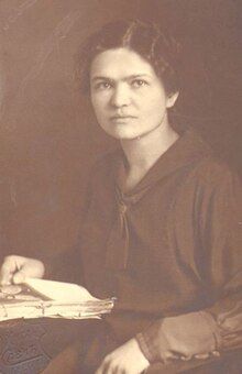 A woman in a sailor suit seated at a desk with her right hand on a manuscript