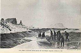 Nine men in dark clothing stand or sit around a small wooden boat that has been dragged on to a rocky shore, with rocks and icy peaks in the background.