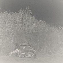 A black-and-white photo of a car parked in front of a hill covered in tall grass.